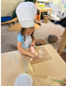 A young girl wearing a chef's hat rolling dough on a sheet of parchment paper